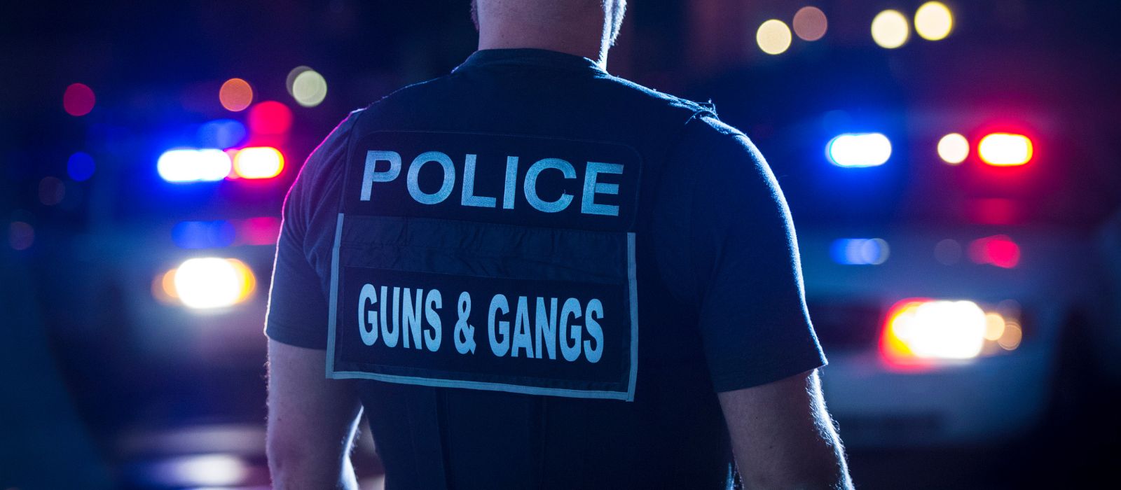 A member of the Guns and Gangs Units stands in front of two police cruisers at night.