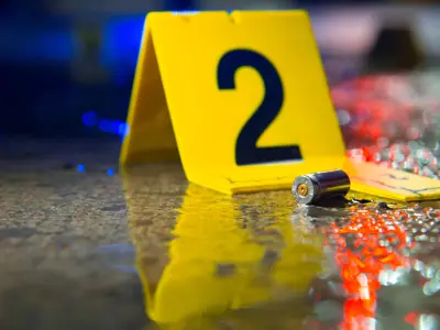 Bullet on the wet pavement, police lights reflecting onto the street with an evidence tags that reads '2'. / Une balle sur la chaussée mouillée, les gyrophares de la police se reflétant dans la rue, avec une étiquette de preuve portant le numéro « 2 ».